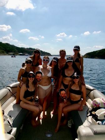 Group of friends on a pontoon boat enjoying a sunny lake day in swimsuits and caps, holding playful face cutouts with a wooded shoreline and blue sky in the background.
