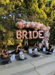 Sunlit backyard bridal yoga with six women in black workout wear seated on mats beneath a rose-gold "BRIDE" balloon sign and pink-and-white balloon arch for a bachelorette celebration.