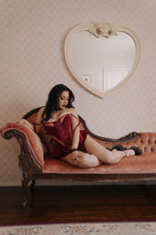 Boudoir portrait of a woman in a red corset reclining on an antique pink velvet chaise lounge under a heart-shaped mirror in a vintage-styled room with patterned wallpaper.