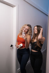 Mirror selfie of two women in a home hallway wearing athleisure—one in an orange crop top holding a phone and a boxed drink, the other in a black crop top and leggings adjusting her hair.
