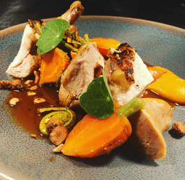 Fine-dining roasted chicken with crispy skin, glazed carrots, fiddlehead ferns, toasted hazelnuts and brown jus, garnished with green nasturtium leaves on a speckled blue plate