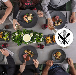 Overhead view of a modern dinner party: people sharing restaurant-style plated gourmet dishes on slate plates — seared steak with mushrooms, prawn with puree, roasted vegetables, asparagus and baby potatoes — red and white wine glasses and a green floral candle centerpiece on a gray table.
