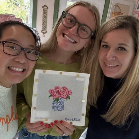 Three smiling women in a cozy craft setting holding a framed cross-stitch of pink roses in a blue patterned vase, cheerful DIY workshop vibe