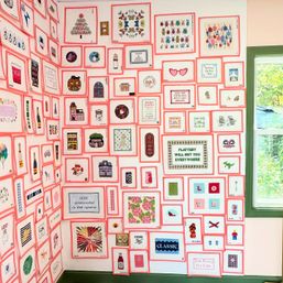 Bright corner wall covered with dozens of small pink‑bordered framed illustrations and sayings arranged in a tight grid beside a green‑trimmed window showing leafy trees — a playful, colorful gallery wall in a sunny interior.