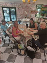 Four women smiling around a floral-covered table in a bright craft studio, doing needlework with colorful thread displays and framed art on checkered tile floors.