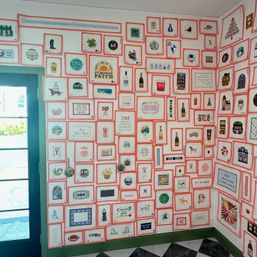 Corner of a colorful shop interior with a teal glass door and black-and-white tile floor, a wall tiled with dozens of small framed prints with pink borders featuring playful illustrations and hand-lettered signs (pumpkin patch, balloons, bottles, houses).