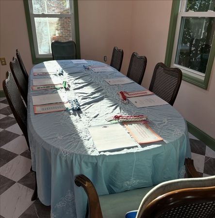 Sunlit dining room with an oval table draped in a light-blue embroidered tablecloth, wooden cane-back chairs, checkered tile floor, and papers with colorful braided cords placed at each seat beside green-trim windows.