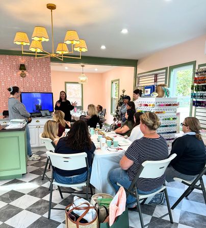 Women in a bright craft studio attending a small-group sewing and embroidery workshop, instructor speaking by a TV, tables with supplies, a multicolored thread wall, pink walls with green trim, and a checkered tile floor.