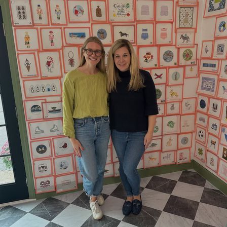 Two smiling women in casual jeans inside a bright boutique-style studio, standing in front of a colorful gallery wall of framed embroidered illustrations with a black-and-white checkered tile floor.