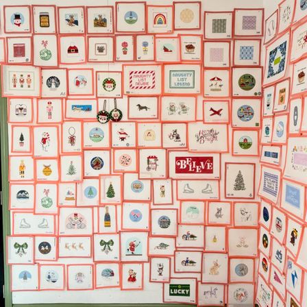 Corner wall covered in a colorful grid of small pink‑bordered holiday greeting card prints — Christmas trees, wreaths, nutcrackers, ornaments and cheerful "Believe" and "Merry & Bright" motifs.