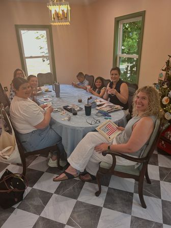 Smiling women and children gathered for a DIY crafting party around a round table in a sunlit home dining room with green-trim windows, checkered tile floor and a decorated holiday tree.