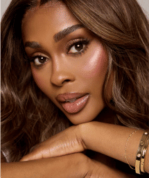 Glam beauty close-up of a woman with glowing skin, shimmery pink blush, glossy nude lips, long lashes and wavy brown hair, chin resting on crossed arms wearing gold bracelets, studio makeup portrait