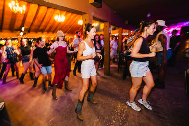 Energetic country line dance in a rustic indoor dance hall, people boot‑scootin' on a wooden floor under warm string lights, many wearing cowboy boots and casual summer outfits