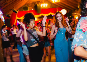 Two women dancing at a lively indoor dance party in a wooden‑beamed venue with colorful lights and a crowded floor