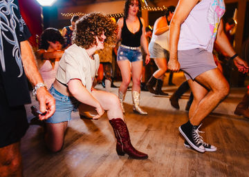 People dancing on a wooden dance floor in a lively country-style indoor venue, wearing denim shorts, cowboy boots and sneakers under string lights