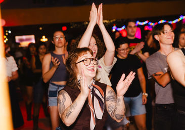 Smiling tattooed person with glasses and a neck scarf clapping in a lively indoor music venue crowd, surrounded by cheering people and colorful string lights — energetic concert/nightlife scene.