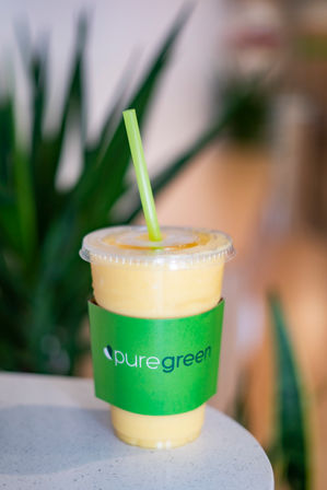 Refreshing yellow mango smoothie in a clear plastic to-go cup with a bright green sleeve and straw, sitting on a cafe table with blurred indoor plants in the background