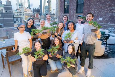 Smiling diverse group on an urban rooftop holding small bonsai trees in a casual team‑building scene with city skyscrapers in the background.