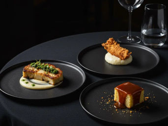 Three fine-dining tasting menu plates on a black tablecloth: seared rectangular protein on a creamy puree with herb oil and microgreens, a crispy breaded strip atop mashed potatoes, and a square sponge cake drizzled in glossy caramel, minimalist elegant presentation with a wine glass in the background.