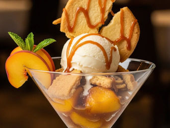 Peach cobbler sundae in a martini glass with a scoop of vanilla ice cream, caramel drizzle, fresh peach slices, crumbled graham crackers, cookie shards and a mint garnish