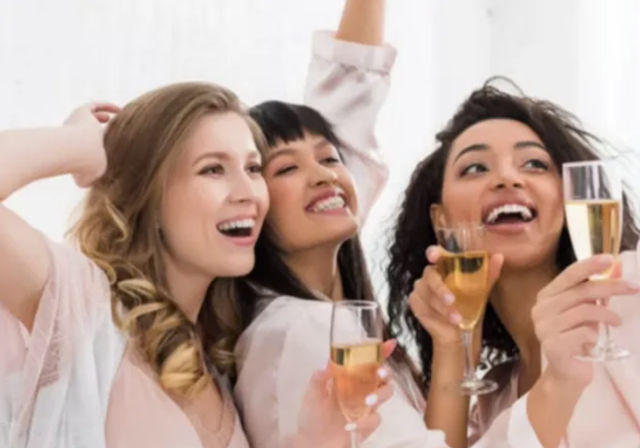 Three smiling women in pastel robes toasting champagne flutes indoors at a joyful bridal shower or bachelorette celebration