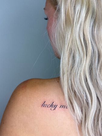 Close-up of an upper back with long blonde hair showing a small, playful script shoulder tattoo that reads 'lucky me'.