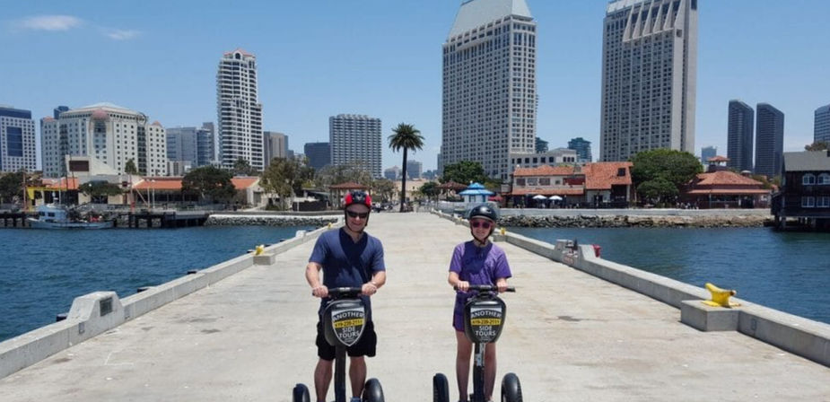 Two helmeted riders on Segways cruise a sunny waterfront pier in San Diego, with palm trees, blue bay water on both sides and a downtown high‑rise skyline in the background.
