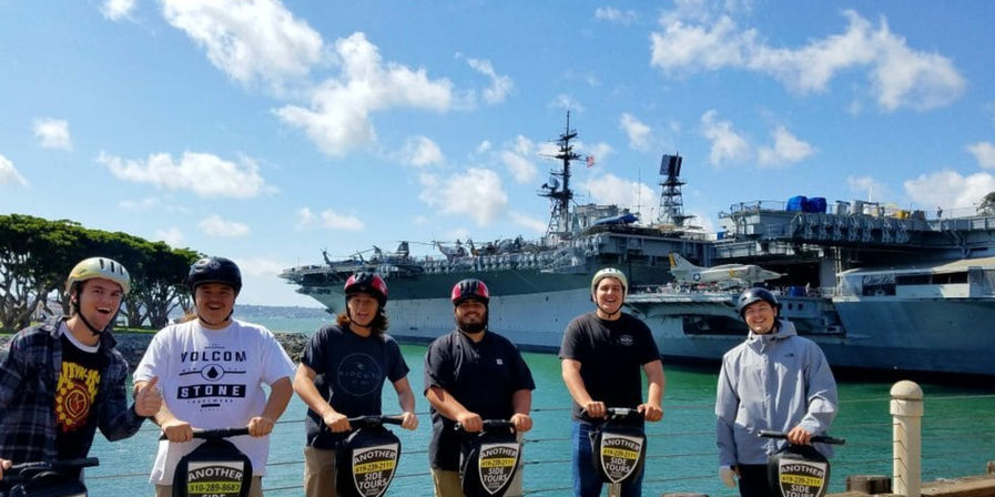 Six people on electric tour scooters along the San Diego waterfront with a large docked aircraft carrier in the background under a sunny blue sky.