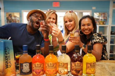 Four adults smiling and raising shot glasses behind a row of colorful spirits bottles in a bright tasting room, celebrating a lively bar-style tasting.