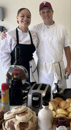 Two smiling chefs in white uniforms behind a breakfast counter with baskets of English muffins and rolls, a toaster and waffle iron, a bottle of maple syrup and jar of jam — cozy local cafe breakfast spread.