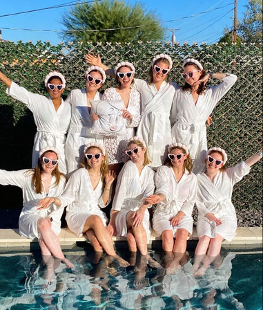 Ten women in white robes and pink heart-shaped sunglasses posing poolside with their feet in the water, playful bachelorette/bridal party at a sunny backyard pool