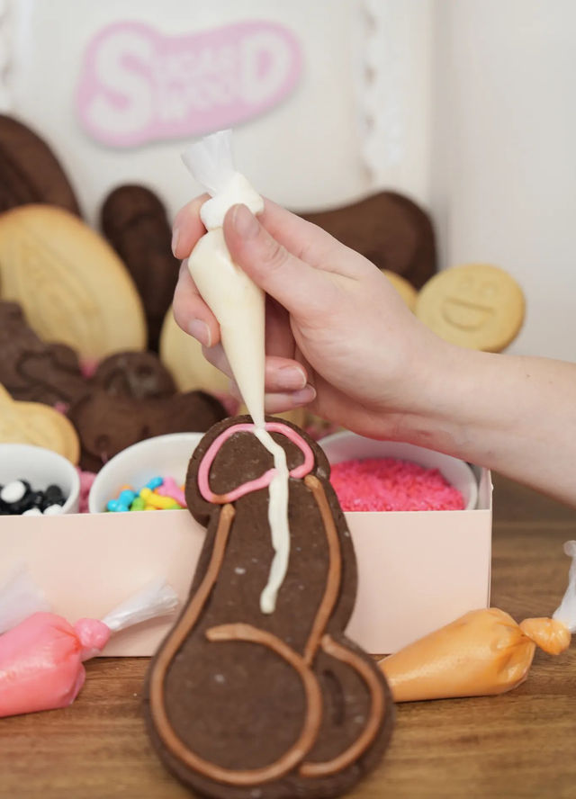 Hand piping white icing onto a chocolate novelty cookie shaped like a penis at a cookie-decorating station on a wooden table with colorful sprinkles and icing bags