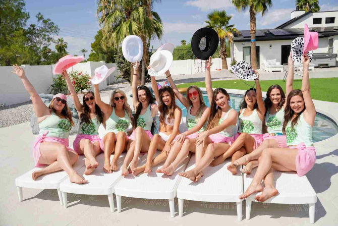 Ten women in matching mint tank tops and pink wraps cheering poolside, waving cowboy hats in a sunny palm-tree backyard beside a modern house.
