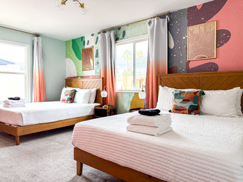 Bright modern guest room with two wooden platform beds, white quilts and folded towels, colorful geometric pillows, coral-ombre curtains, abstract mural accent wall and window with palm trees