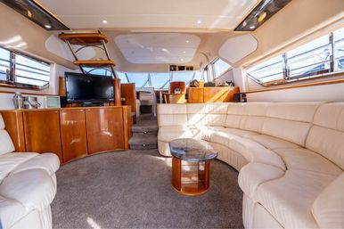 Sunlit luxury yacht salon with cream leather wraparound seating, glossy wood cabinetry, glass-topped coffee table, flat-screen TV, carpeted floor and large windows overlooking the marina.