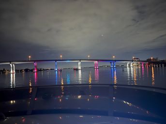 Nighttime waterfront bridge with pink, blue and white LED-lit arches reflecting on calm bay, view from a boat under a cloudy sky