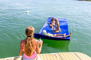 Four friends relaxing on a bright blue floating mat tied to a boat on a sunny lake — two sitting, one reclining on the mat, and one on the boat deck holding a cold drink.