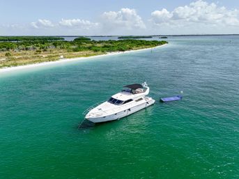 White motor yacht anchored in turquoise coastal bay near a sandy beach and green mangrove islands, with a purple swim mat floating beside the boat under a partly cloudy sky.