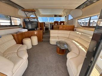 Sunlit luxury yacht salon with cream leather curved sofas, marble-top coffee table, glossy wood cabinetry and TV, large windows with marina views and steps up to the forward helm