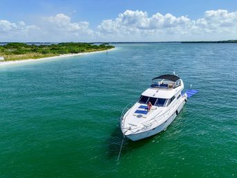 Luxury white yacht anchored in clear turquoise coastal waters off a sandy shoreline, a person relaxing on the bow under a sunny blue sky with puffy clouds.