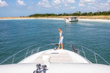 Woman standing on the bow of a yacht in turquoise water near a sandy island beach with an anchored tour boat on a sunny day