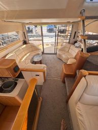 Sunlit luxury yacht salon with curved beige leather sofas, marble-top coffee table, carpeted floor, spiral stainless-steel staircase and sliding glass doors opening to a teak aft deck and marina view.