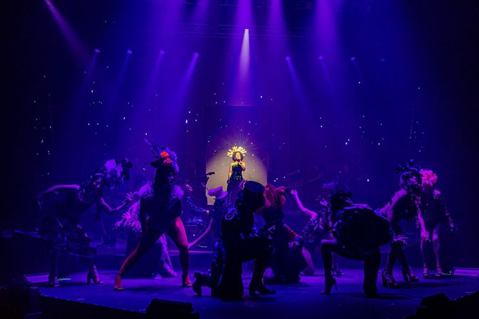 Lead singer in a glowing sunburst headdress stands in a bright spotlight on a theater stage, surrounded by costumed dancers striking dramatic poses under vibrant purple lighting during a lively cabaret-style live performance.