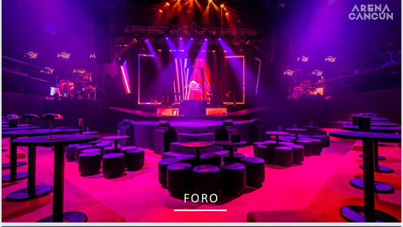 Empty indoor concert venue interior in Cancún with a central illuminated stage, large LED screens and purple-pink spotlights, round low cocktail tables and pouf seating on a red floor.