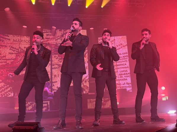Four male singers in black outfits holding microphones, performing on an indoor stage bathed in pink and yellow lights with a map-patterned video backdrop