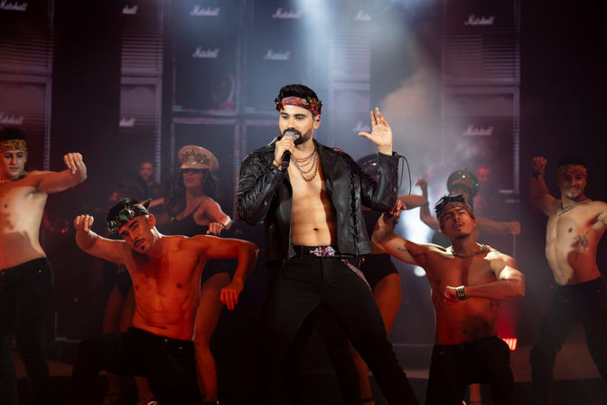 Energetic male singer in an open leather jacket and headband performs on a smoky live concert stage with shirtless backup dancers and dramatic lighting