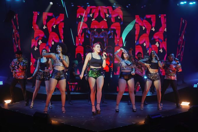High-energy live concert with a group of singers and dancers in sparkly and leather outfits performing a choreographed routine on a lit stage with vibrant red-patterned LED screens and dynamic stage lighting.