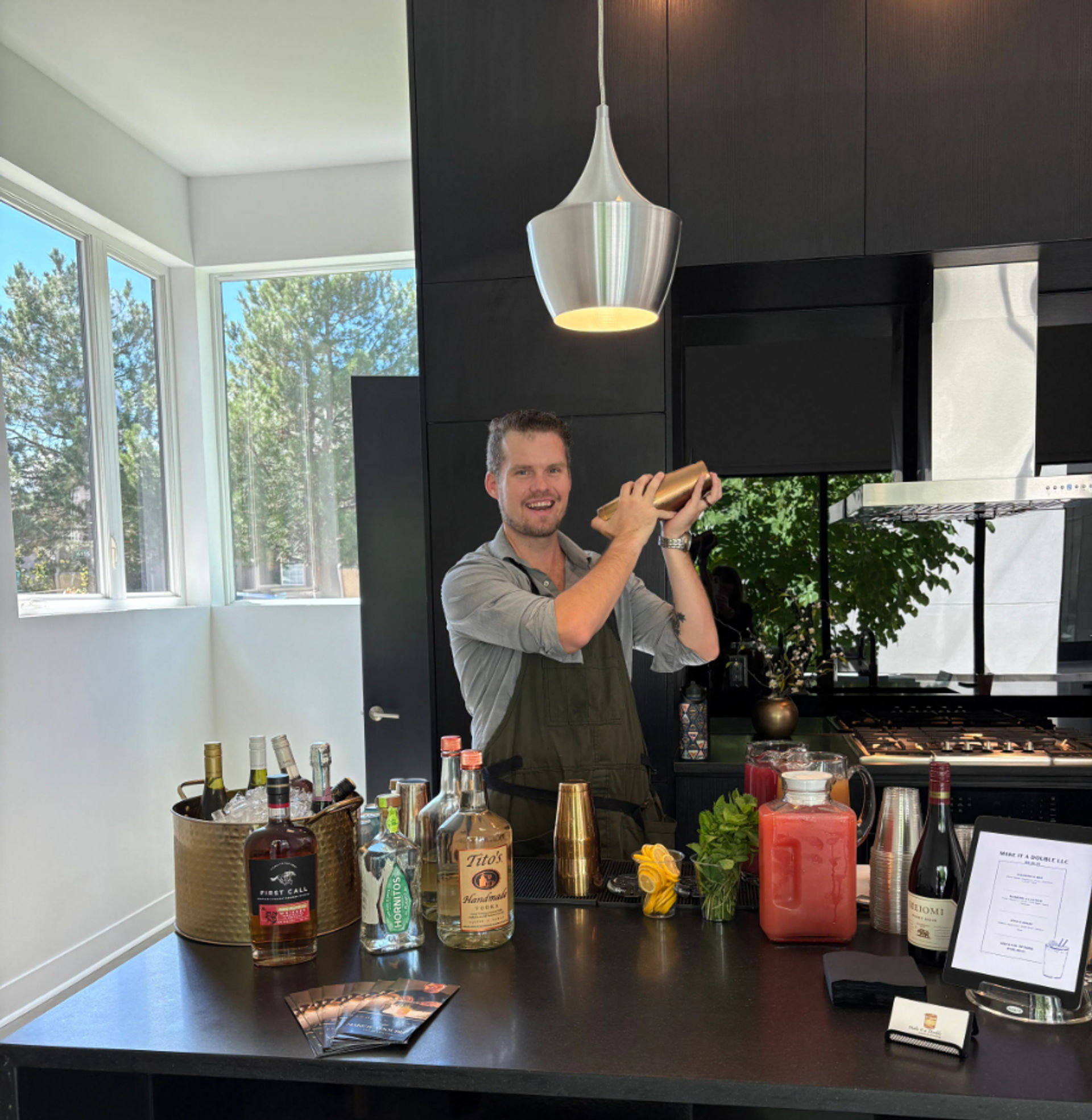 Smiling bartender in a modern sunlit kitchen shaking a cocktail at a home bar with liquor bottles, garnishes, pitchers and large windows overlooking pine trees.