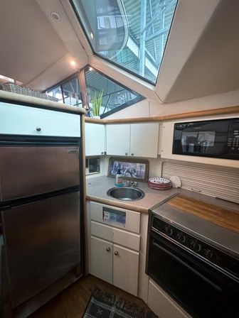 Compact yacht galley with stainless refrigerator, built-in microwave and stove, round sink on a laminate counter, and large angular skylight showing marina structure above