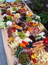 Vibrant outdoor charcuterie grazing board on a paper-lined table in a garden — assorted cheeses, cured meats, crackers, grapes, strawberries, dried apricots, olives, cornichons, nuts, mini peppers, cucumbers and dips.